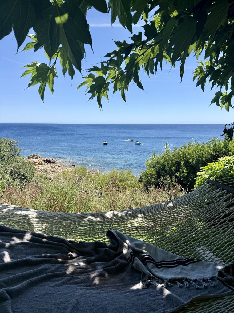 A hammock and a striped towel in dappled shade, the Mediterranean and two moored boats below