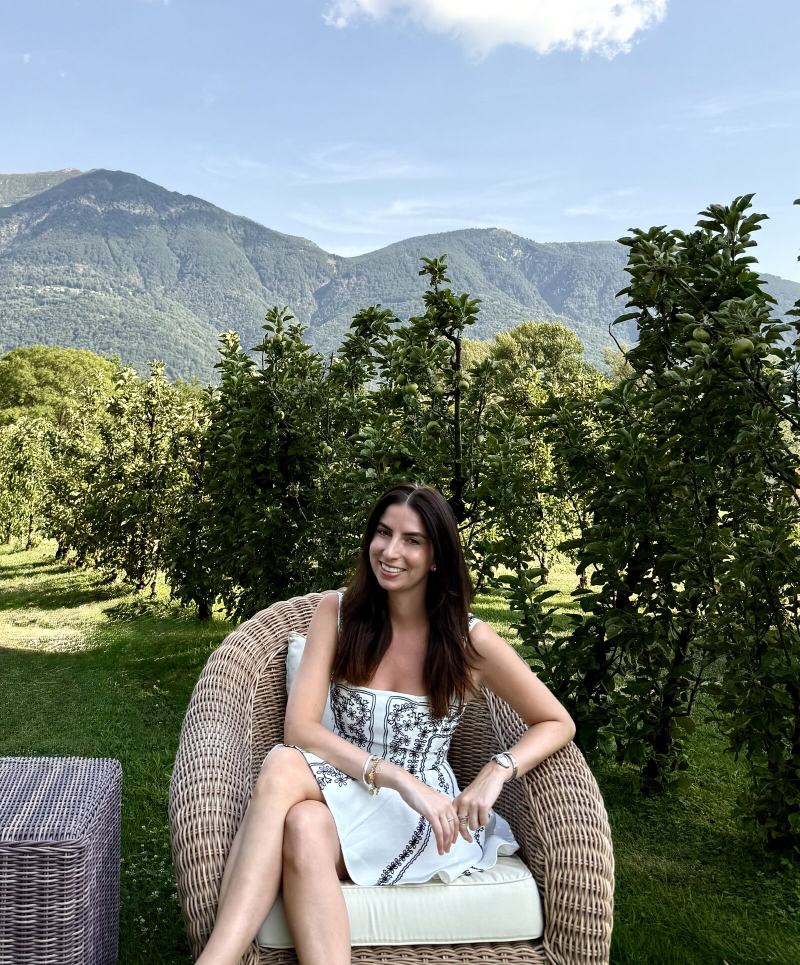Anna Kovarbasic, founder and host of Entre Nous, seated in a wicker chair in an orchard with mountains behind her