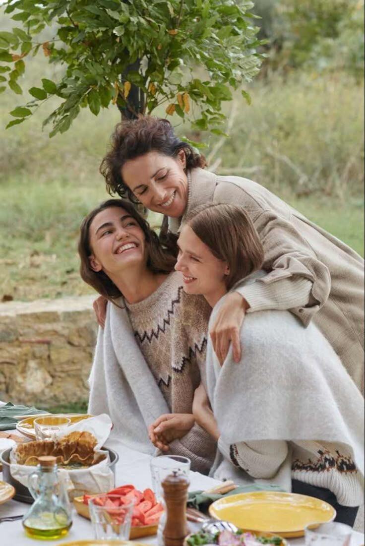 A mother and her two daughters laughing together, wrapped in blankets at a garden picnic