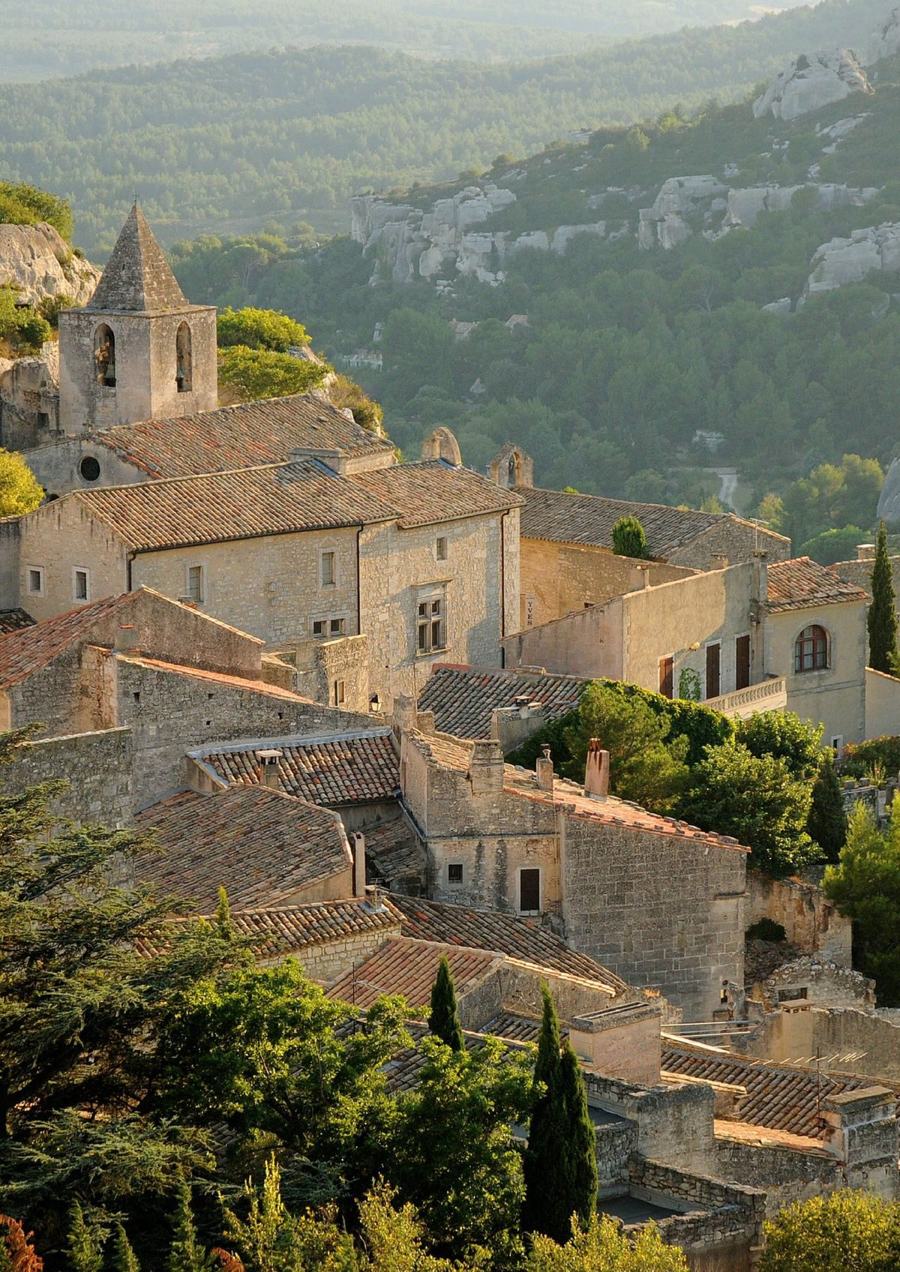 A Provençal hill village of pale stone and tiled roofs in golden evening light, the Alpilles rising behind