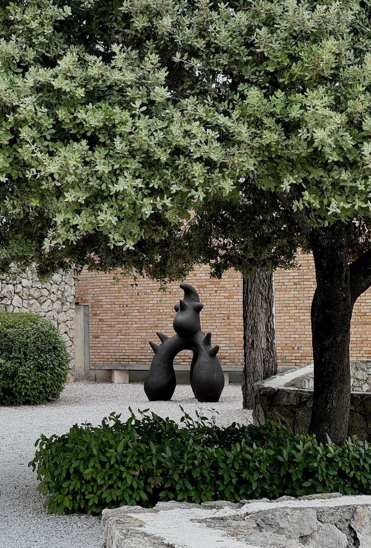 A dark ceramic sculpture standing on gravel beneath an olive tree, clipped hedges and old brick and stone walls behind