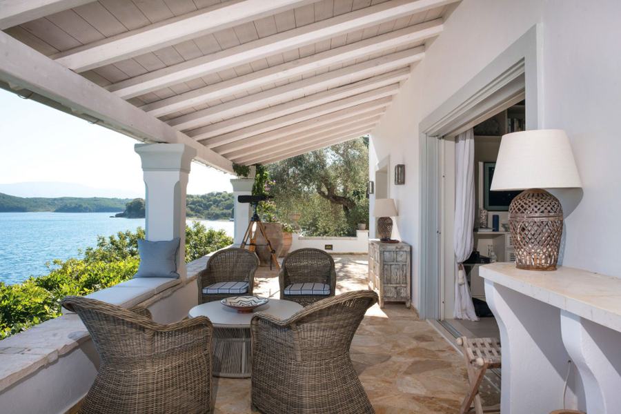 A covered stone terrace with wicker armchairs and a cushioned bench, looking out over the water through olive branches