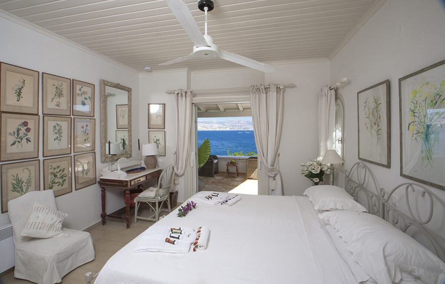A white bedroom with framed botanical prints and doors open to a terrace above the sea